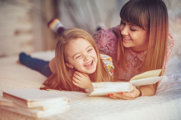 Portrait of a smiling young cute mother and daughter reading a book lying and relax in the bed in a bright big white room